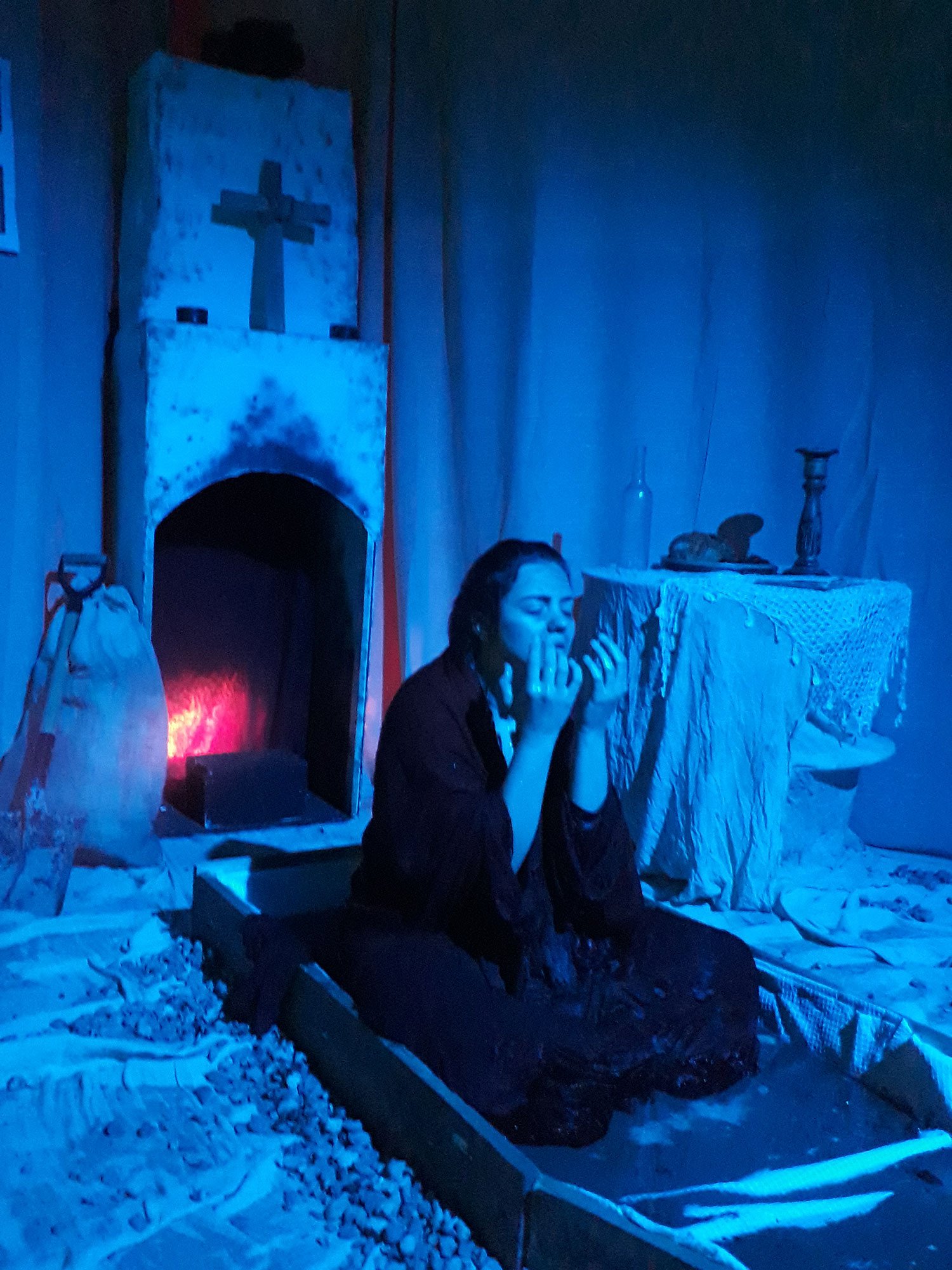 An actor is stooped in prayer, in a boat-There's a fireplace and crucifix behind her